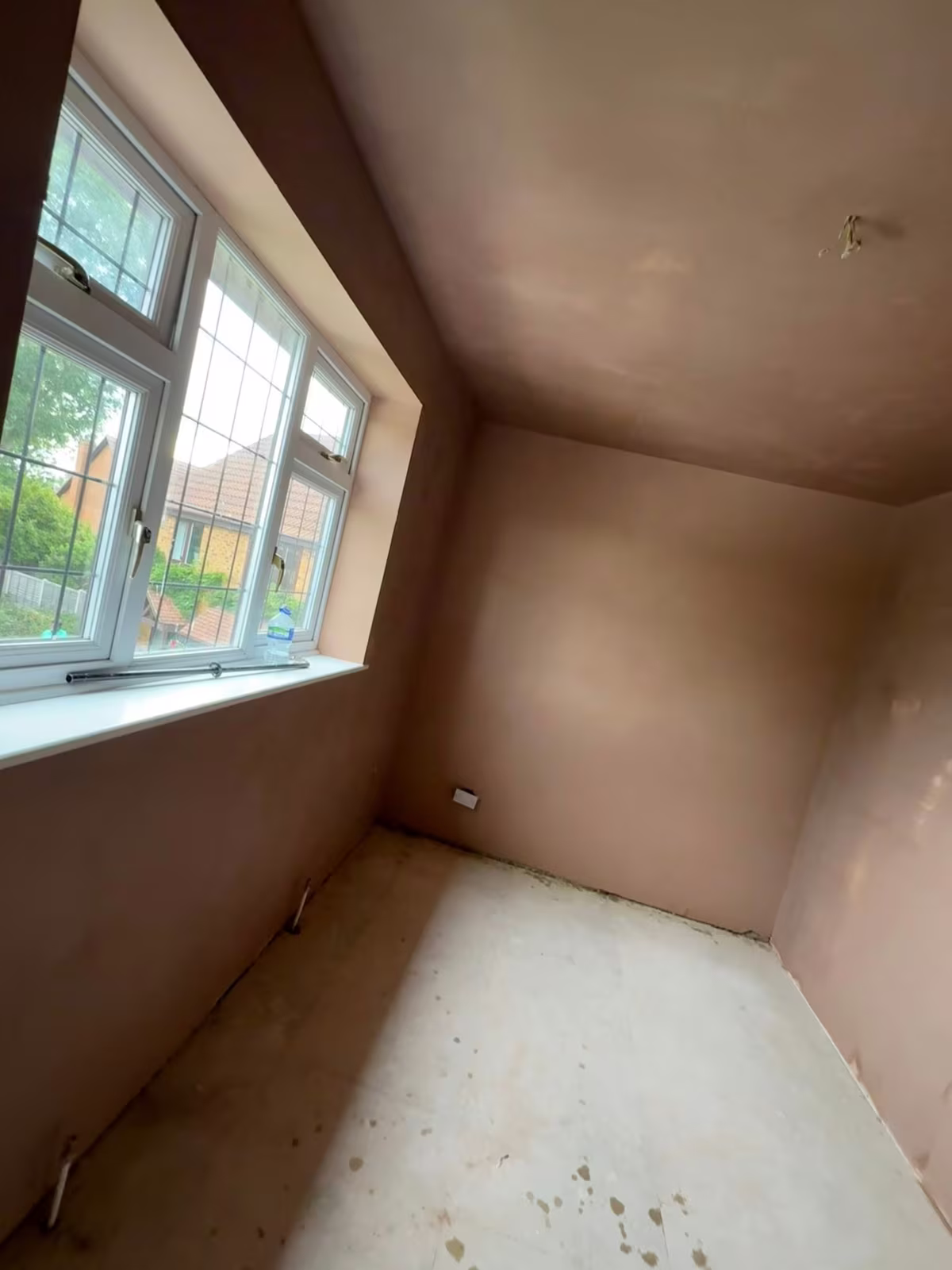 A bedroom with walls and ceiling newly skimmed, still drying pink, and a bay window onto the street